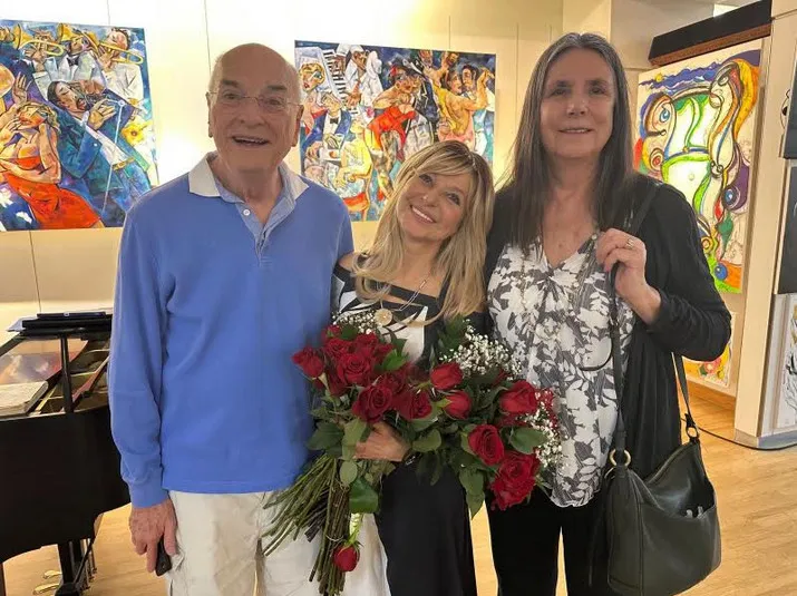 Members with a bouquet of red roses beside the piano, bright paintings on display behind them.